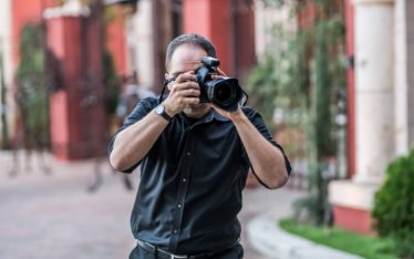 A man in a black shirt stands on a paved street, holding a camera up to his face while taking photos at a corporate event. The background is blurred, showing red buildings and greenery.