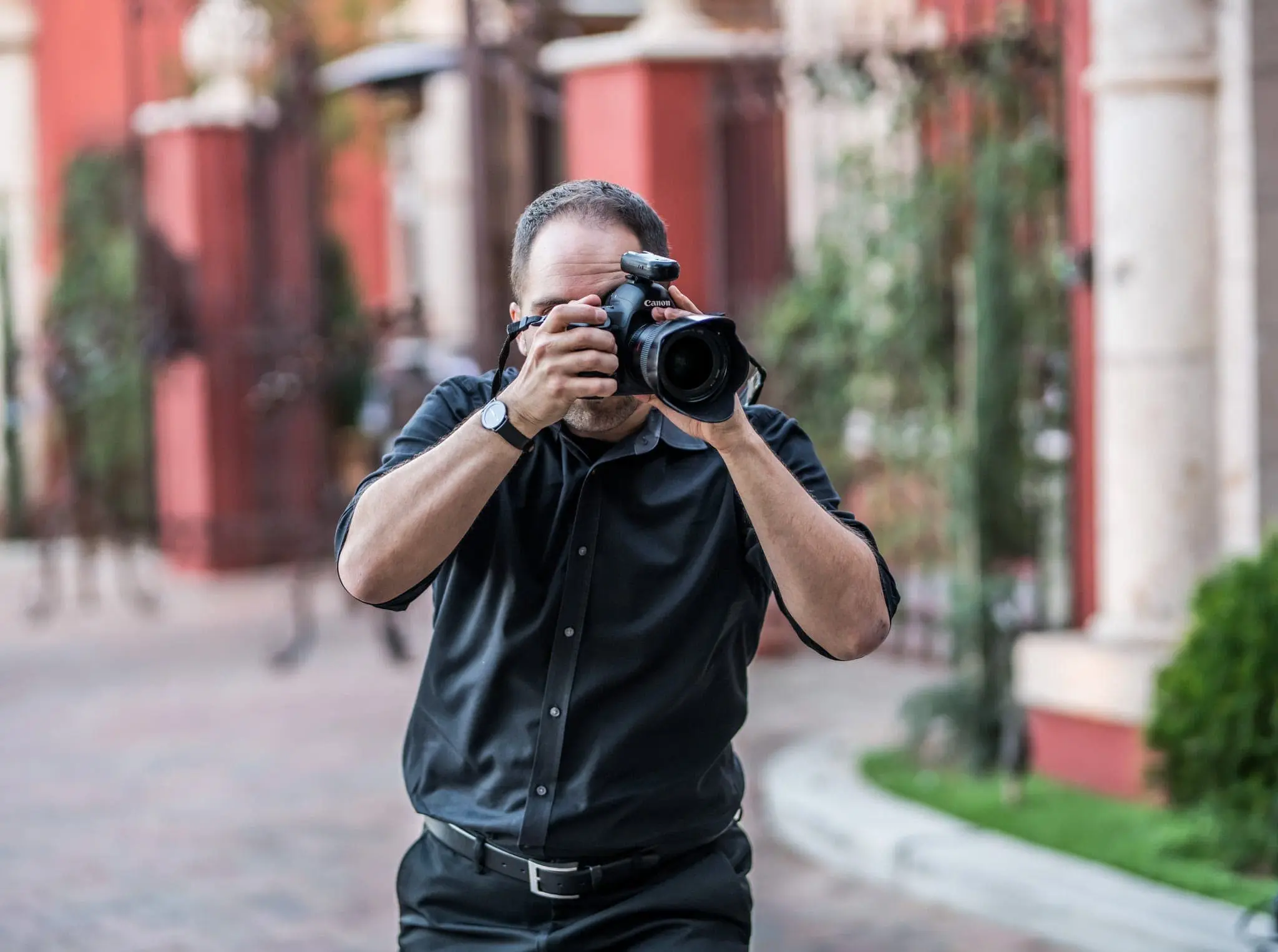A man in a black shirt stands on a paved street, holding a camera up to his face while taking photos at a corporate event. The background is blurred, showing red buildings and greenery.