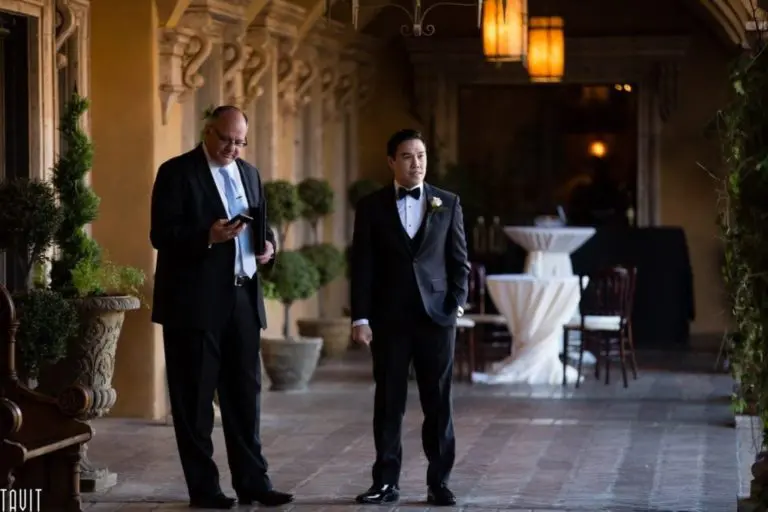 Two men in formal attire stand in a warmly lit, elegant hallway, capturing the essence of corporate event photography. One man is looking at a tablet, while the other, wearing a tuxedo, stands with his hands by his sides. Decorative plants and tables with white tablecloths are visible in the background.