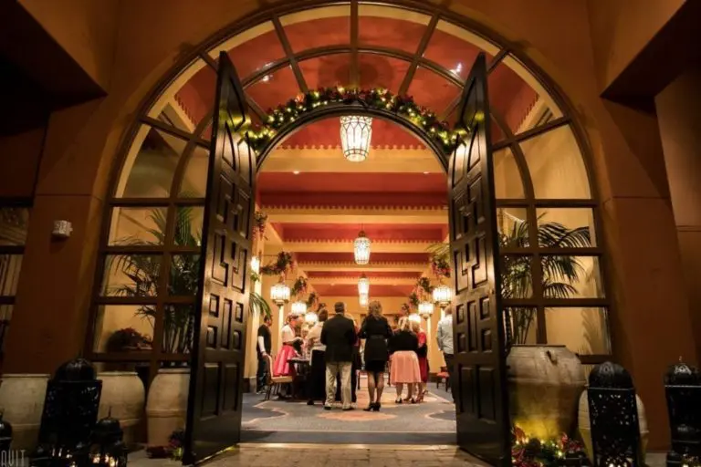 Elegant entrance with large open doors leading to a warmly lit interior decorated with plants and festive garlands. A group of people, dressed in formal attire, are gathered inside for a corporate event, creating a welcoming atmosphere perfect for event photography.