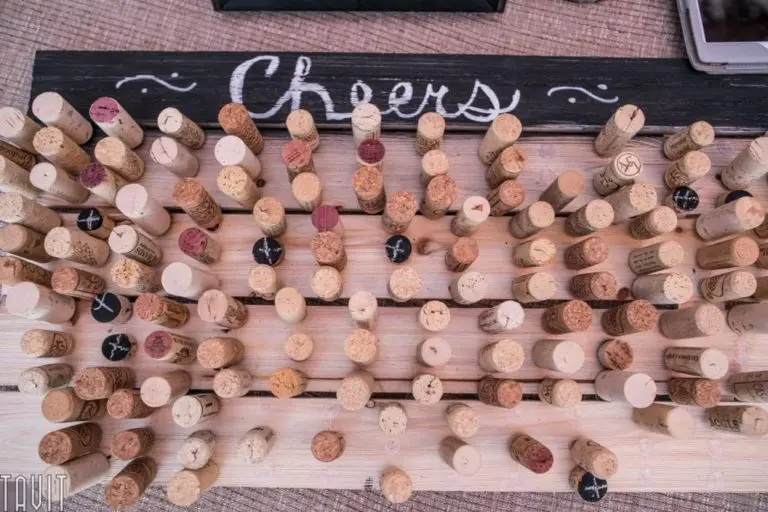A collection of wine corks arranged vertically on a wooden board serves as an eye-catching backdrop for corporate event photography, with the word "Cheers" elegantly painted in white across a sleek black background at the top.