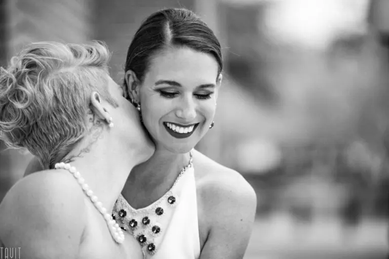 A black-and-white photo of two women dressed elegantly at a corporate event; one with short hair and a pearl necklace kisses the other, who is smiling brightly and wearing a jeweled neckline.