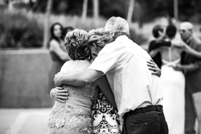 Three adults embrace in a group hug outdoors at a corporate event, their faces partially hidden. In the blurred background, others are hugging or dancing. The image is in black and white.