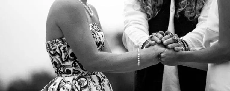 Two people holding hands during a ceremony, with an officiant standing behind them. One wears a patterned dress, the other a sleeveless white outfit. The black-and-white image evokes the formality of a seminar or corporate event.