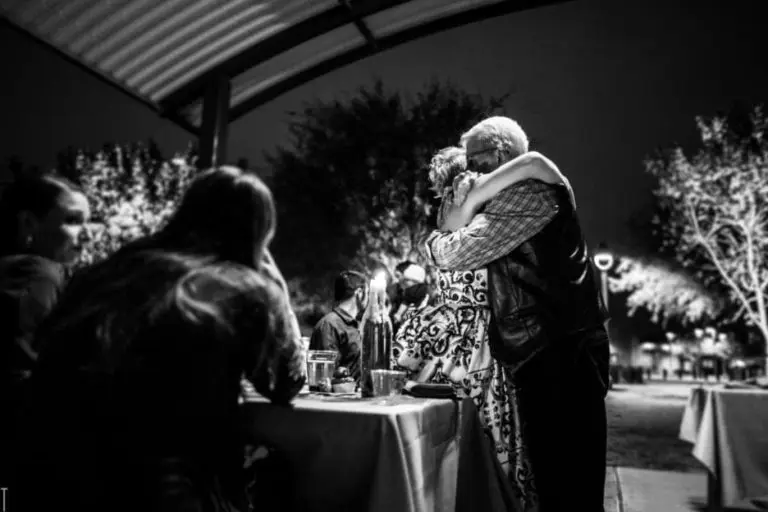 A black-and-white photo shows two people hugging warmly under a pavilion at night during a corporate event, while others sit at tables nearby. Trees decorated with lights are visible in the background.
