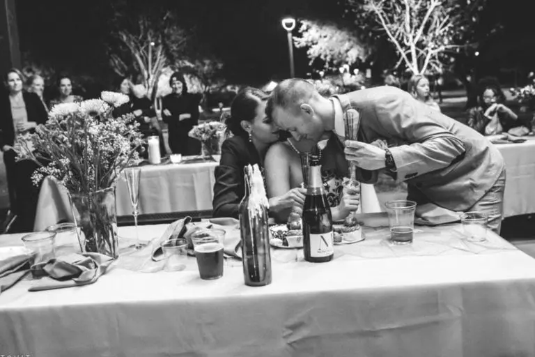 A couple shares a kiss while seated at a decorated table during an outdoor evening corporate event, with guests and trees in the background. The scene is captured in elegant black and white.