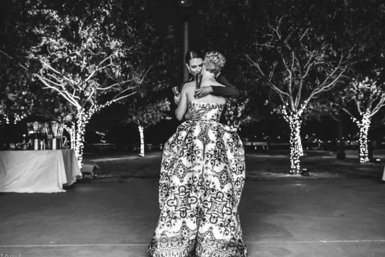 Two women in formal dresses hug while slow dancing outdoors at night during a corporate event. Trees wrapped in string lights glow in the background, creating an intimate, romantic atmosphere. The image is in black and white.