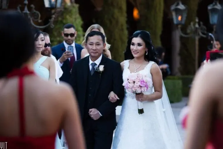 A bride in a white gown holds a bouquet of pink and white roses, walking arm-in-arm with a man in a black suit. They are outdoors at an event surrounded by dressed-up guests, some engaged in event photography. Trees and lampposts provide the perfect backdrop for this memorable scene.