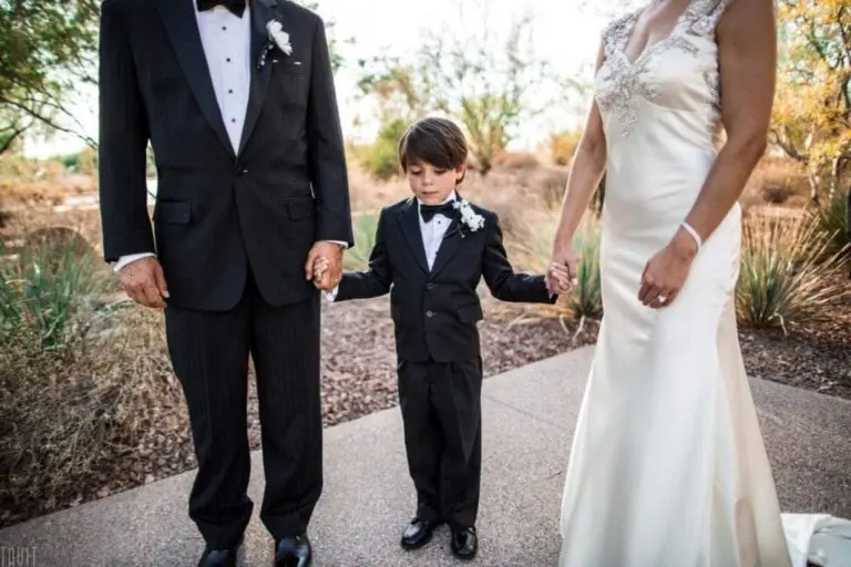 A young boy in a tuxedo holds hands with a man in a black suit and a woman in a white wedding dress. Captured brilliantly through event photography, they stand on a pathway outdoors, surrounded by lush greenery.