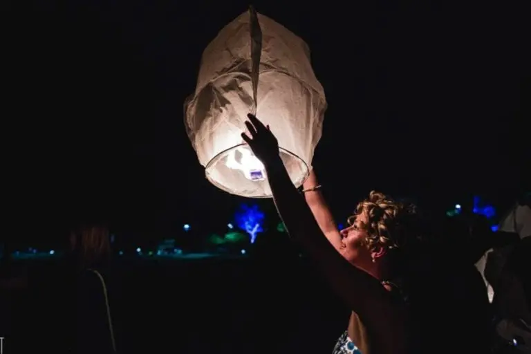 At a corporate event, a woman holds and releases a glowing paper lantern into the night sky, her face illuminated by its light against a dark background dotted with faint, colorful lights.
