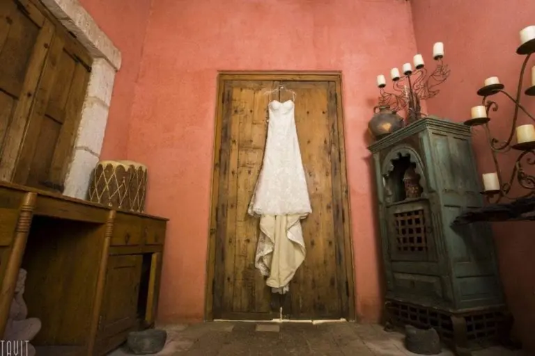 A white lace wedding dress hangs on a wooden door in a rustic room with red walls, wooden furniture, a drum, and a blue cabinet—an elegant contrast to the usual corporate event or seminar setting.