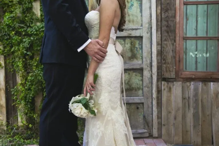 A bride in a lace wedding dress holding a bouquet of white roses stands close to a groom in a dark suit, outdoors by a rustic wooden wall and green vines—far from any seminar or corporate event.