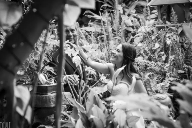 A bride in a white dress smiles and affectionately touches an older man's face during a corporate event, surrounded by lush greenery in an outdoor garden setting. The photo is black and white.