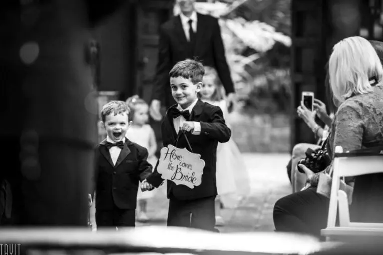 Two young boys in tuxedos walk down an aisle at a wedding seminar, smiling and holding a sign that reads "Here comes the bride," while guests watch and take photos.