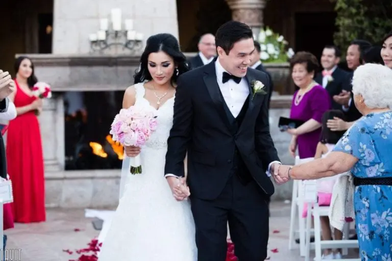 A bride in a white gown holds pink flowers, walking hand-in-hand with a groom in a black tuxedo. They're smiling and passing guests, as if cameras are capturing every moment of this special event, with a lit fireplace casting a warm glow in the background.
