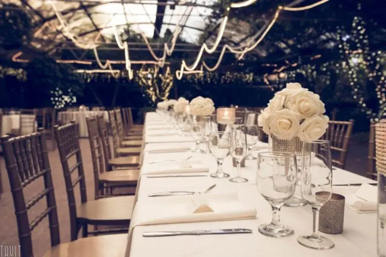 A long, elegant banquet table is set for a corporate event with white cloth, glassware, and napkins, decorated with white rose centerpieces and surrounded by wooden chairs. String lights and greenery create a warm, romantic atmosphere.