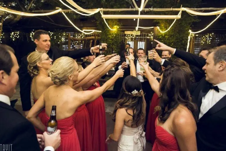 A group of people in formal attire, with women in red dresses and men in black suits, stand in a circle raising their glasses for a toast at an outdoor corporate event decorated with string lights.