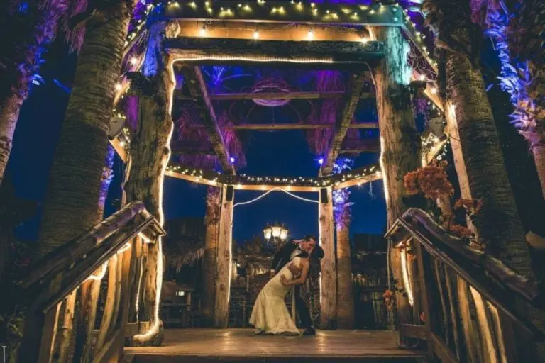 A couple in wedding attire shares a romantic dip and kiss under a rustic, wooden gazebo—typically used for seminars or corporate events—decorated with string lights at night, surrounded by palm trees and soft purple lighting.