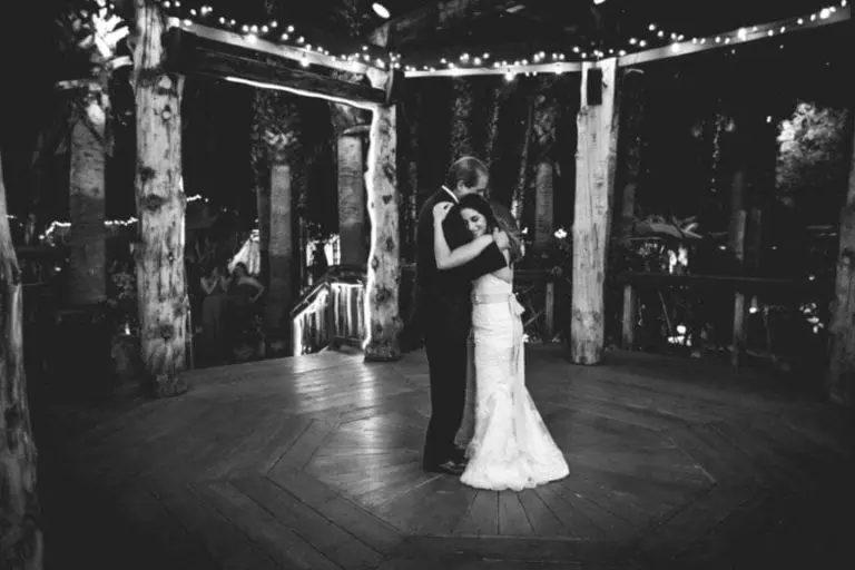 A bride and groom share a quiet first dance on a wooden outdoor deck under string lights, surrounded by tall trees at night. The scene is in black and white, evoking the intimacy of a private moment rather than a seminar or corporate event.