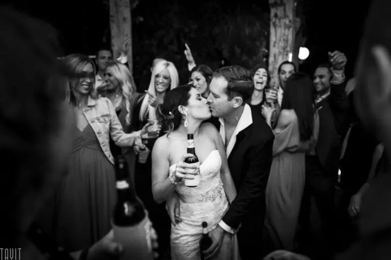 A bride and groom share a kiss at their wedding reception, surrounded by smiling friends and guests—some recalling moments from a recent seminar—in a lively, celebratory atmosphere. The photo is in black and white.