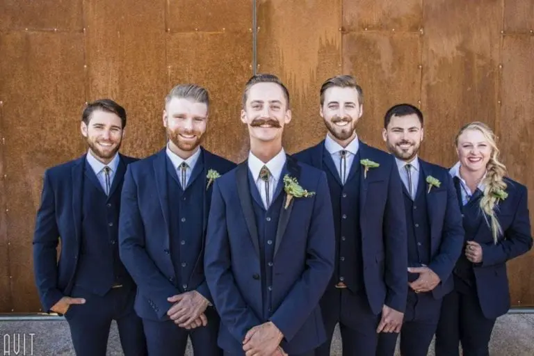 Six people dressed in matching navy blue suits and ties stand smiling in front of a rust-colored wall. Five men and one woman, all wearing boutonnieres, pose together at a festive corporate event in a formal setting.