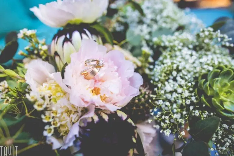 A pair of wedding rings rests on a pale pink peony, surrounded by green leaves, white baby's breath, and other floral arrangements—elegant details that could enhance the ambiance of a seminar or corporate event in soft, natural light.