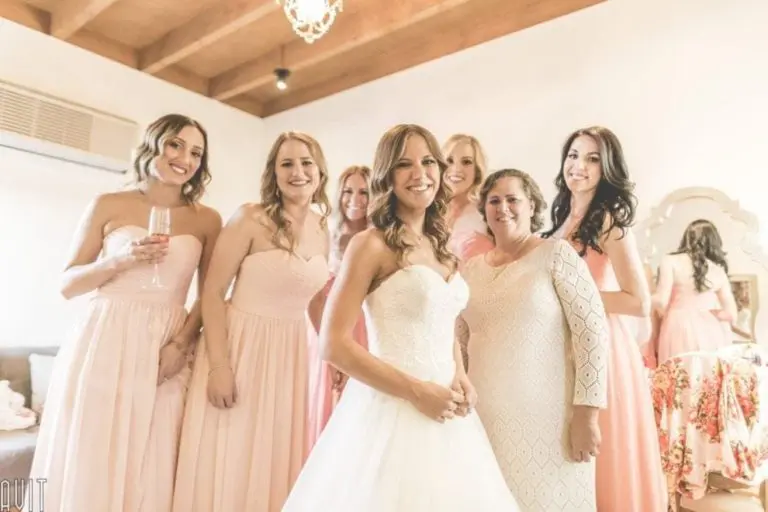 A bride in a white strapless gown stands smiling with her bridesmaids, who wear blush pink dresses, and one woman in a cream lace dress, all posing together in a bright, elegant room set up for a seminar or corporate event.
