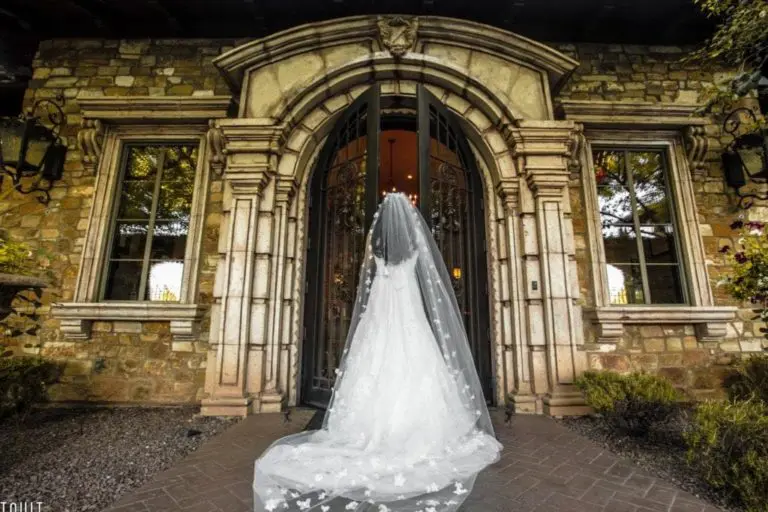 A bride in a flowing white gown and veil stands before grand arched wooden doors surrounded by a stone facade, reminiscent of an elegant corporate event. She faces the entrance, partially obscured by her train embellished with floral designs, as if captured in exquisite event photography.