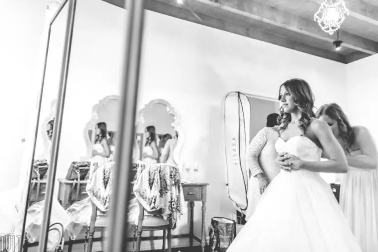 A bride in a strapless wedding dress stands while two women help her get ready in a bright, elegant room with mirrors and vintage furniture—far from any seminar or corporate event. The scene, reflected in a mirror, is captured in black and white.