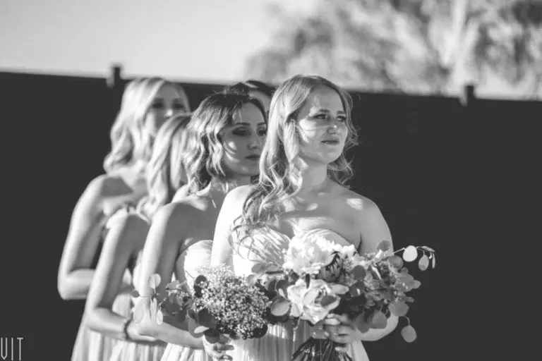 A group of bridesmaids in light dresses stand in a line outdoors, each holding a bouquet of flowers. The black-and-white image evokes the poised unity often seen at a seminar, with the women looking ahead with serene expressions.
