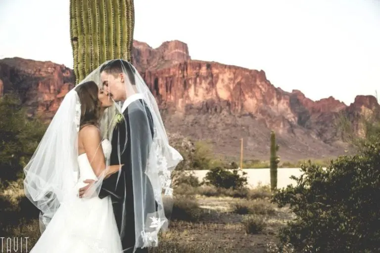 A bride and groom stand close together under a veil, embracing in front of a large cactus at sunset—the scene contrasts the natural beauty with the nearby venue where a seminar or corporate event has just wrapped up.