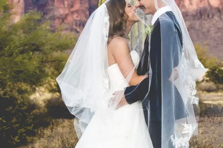 A bride and groom in wedding attire stand close together, smiling under a shared veil in a desert landscape, as if taking a break from a seminar, with a large cactus and rocky mountains in the background.