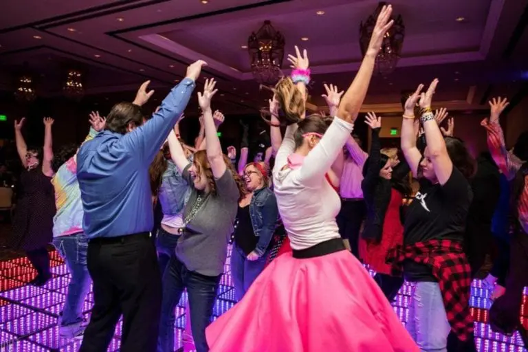 A group of people dance enthusiastically on a colorful, illuminated floor at a lively corporate event, raising their arms and smiling under chandeliers in a decorated banquet hall.