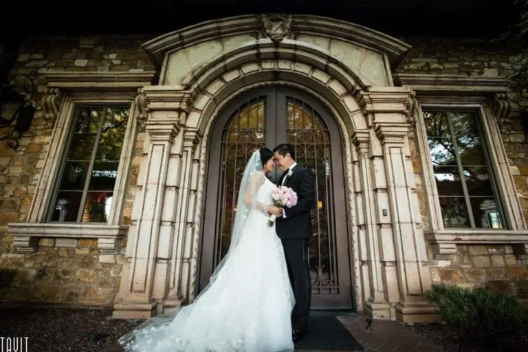 A bride and groom share a kiss in front of an ornate, arched doorway with intricate stonework, capturing the essence of event photography. The bride holds a bouquet of pink and white flowers. The building's rustic facade adds a romantic backdrop to the scene.