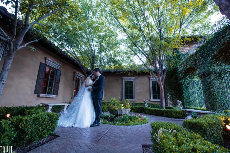 A couple in wedding attire embraces on a brick path in a lush, green courtyard. The bride wears a long white gown, and the groom is in a dark suit. Surrounded by tall trees and manicured hedges, it's an event photography dream against the sunlit walls of a rustic building.