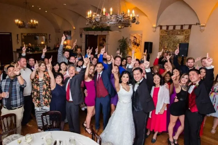 A large group of people celebrating at a wedding reception in a decorated room. The bride and groom, in the center, are surrounded by guests, some raising their hands joyfully. Capturing the essence of event photography, tables with plates and glasses are visible under elegant chandeliers.