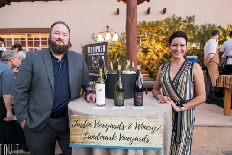 A man in a suit and a woman in a striped dress stand beside a table adorned with wine bottles, capturing the essence of event photography. A sign reads "Justin Vineyards & Winery, Landmark Vineyards" as people savor the corporate event unfolding in the background.