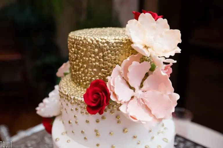 A two-tiered cake, perfect for a corporate event, is decorated with gold pearls. The top tier is adorned with pink and red roses. It features intricate details and a mix of textures, boasting a patterned lower tier and a smooth upper tier.