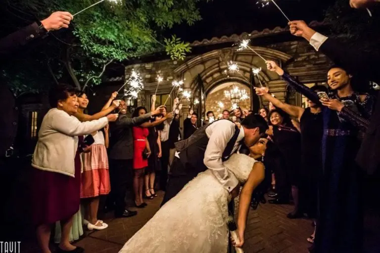 A bride and groom share a romantic kiss while surrounded by guests holding sparklers, capturing the essence of event photography. The couple stands at the center of a stone path under a warm, illuminated entrance, with the scene filled with joyful celebration and evening elegance.