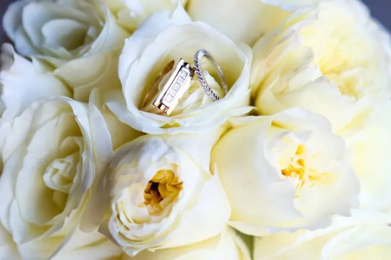 A bouquet of white roses, perfect for a corporate event, with two rings nestled on top. One ring is gold with a small diamond; the other is a thin, silver band. The roses are fresh, with soft petals and a creamy hue.