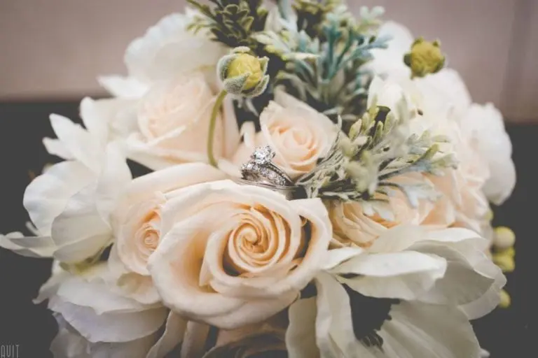 A bouquet of cream-colored roses and greenery, ideal for a corporate event, is adorned with a set of intertwined wedding rings in the center, one boasting a prominent diamond.