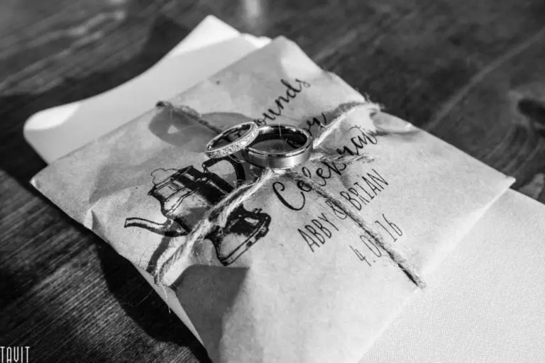 A black and white image captures wedding rings resting on a brown paper envelope tied with twine. Featuring an illustration of a coffeepot, the envelope is labeled "Abby & Brian, 4.10.16." This exquisite example of event photography unfolds against a rustic wooden backdrop.