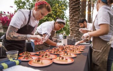 Chefs, wearing aprons and gloves, are meticulously plating multiple dishes on a long table outdoors. As one chef adds an extra ingredient, the scene captures the essence of event photography at its finest. Rows of identical red plates being arranged by three chefs under palm trees enhance this corporate event's ambiance.