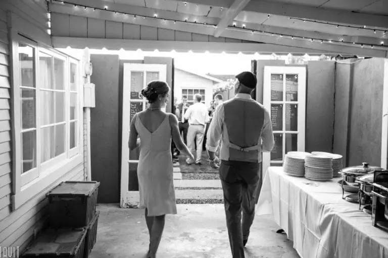 A couple walks hand in hand through a doorway into an outdoor area, dressed in formal attire. The room, captured beautifully through event photography, has string lights, windows, and a long table with stacked plates to the right. Other people are visible in the background. Black and white photo.