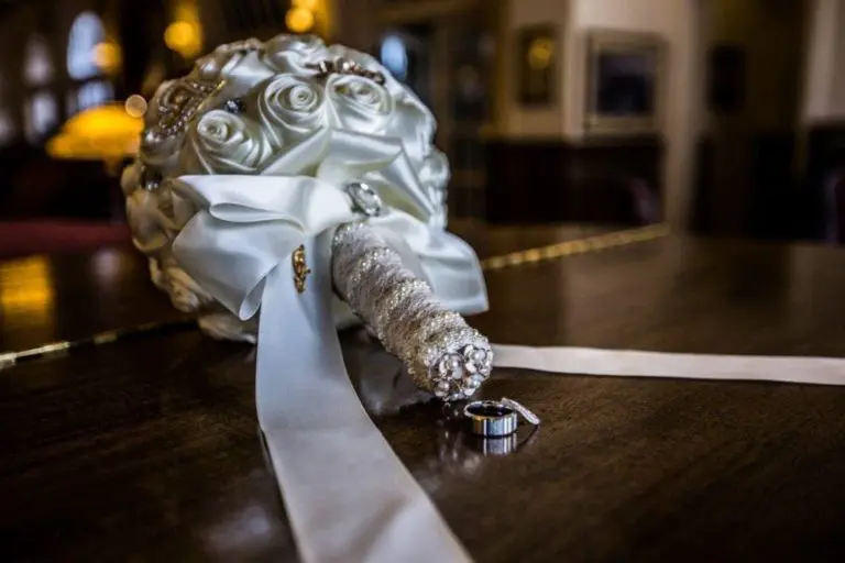 A bridal bouquet of white satin roses, adorned with pearls and tied with a ribbon, rests on a wooden surface. Beside it lie two rings. The background is softly blurred, reminiscent of an elegant indoor setting captured by expert event photography.