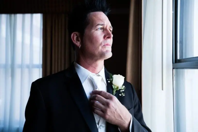 A man in a black suit with a white rose boutonniere stands by a window at a corporate event, adjusting his tie and gazing thoughtfully outside. Sunlight softly illuminates his face and the room behind him.