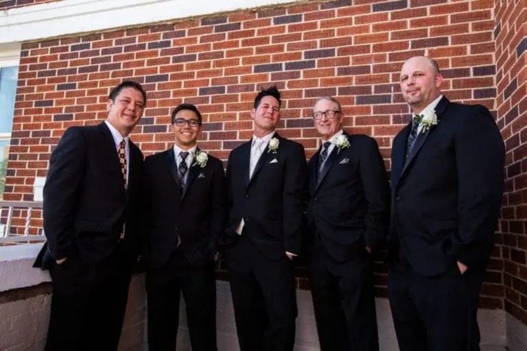 Five men in black suits with ties and boutonnieres stand together smiling in front of a red brick wall, suggesting a wedding, corporate event, or formal seminar.