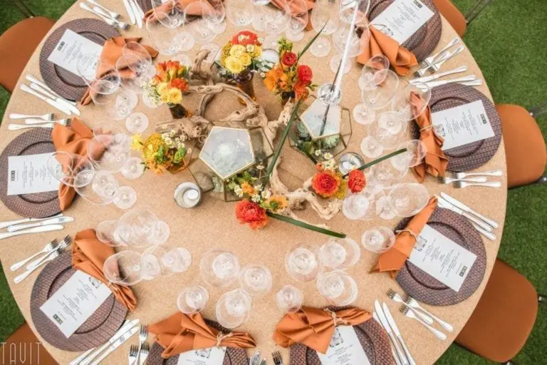 A round dining table set for a corporate event exudes elegance. It features ornate centerpieces with vibrant flowers and decorative elements. Each place setting has a stylishly folded orange napkin, a menu, glasses, and silverware on a textured beige tablecloth. Perfect for event photography.