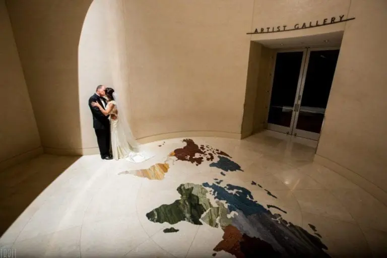 A bride and groom embrace under a spotlight near an entrance labeled "Artist Gallery," standing on a floor map of the world in a spacious, elegantly curved hallway that could host a seminar or corporate event.