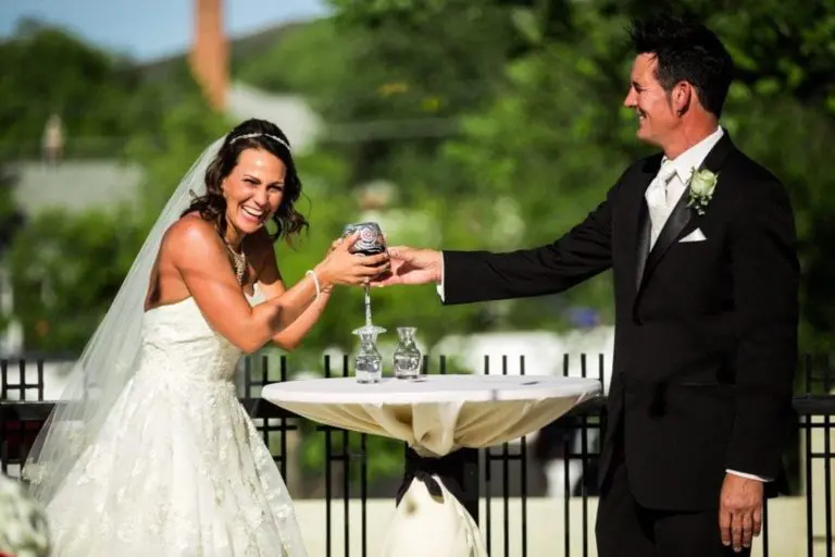 A bride and groom stand outdoors at a small table, laughing as the bride playfully pulls a cup from the groom's hand during their wedding ceremony—not your typical corporate event, but a joyful moment to remember.